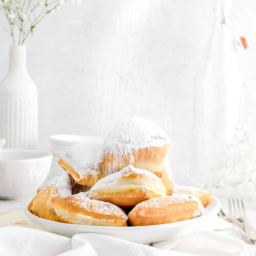close up of stacked beignets being dusted with powdered sugar in a white bowl