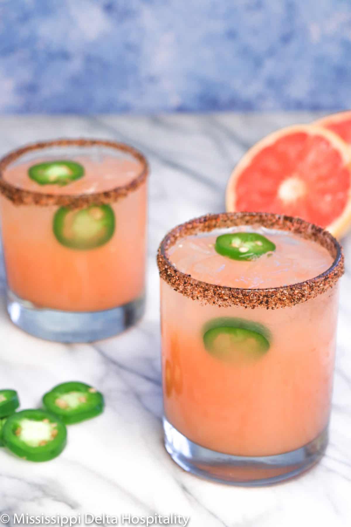 two rocks glasses with spicy grapefruit margarita in them and a chili salted rim on a marble board with jalapeño slices and a grapefruit sliced in half.