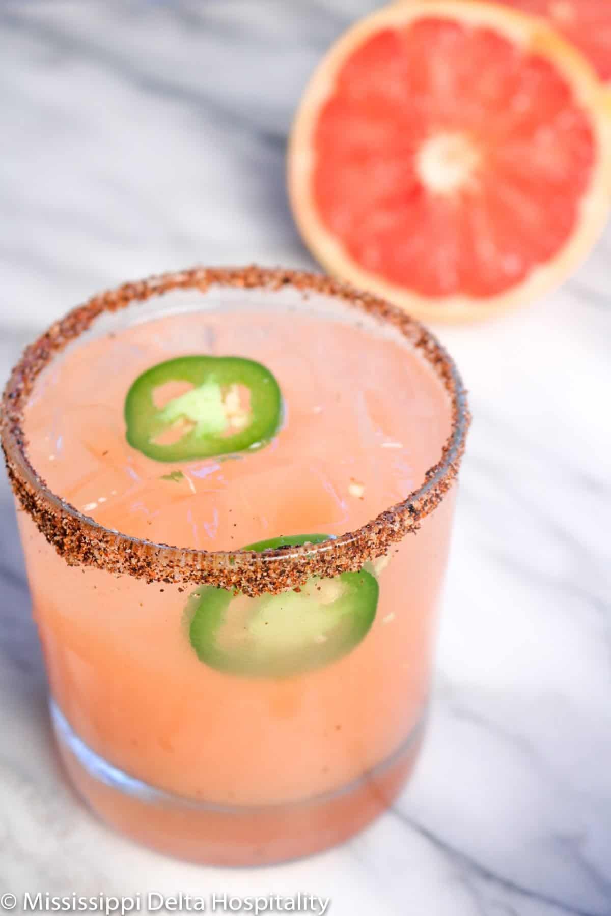 one rocks glass with spicy grapefruit margarita with jalapeño slices on a marble board and a grapefruit sliced in half.