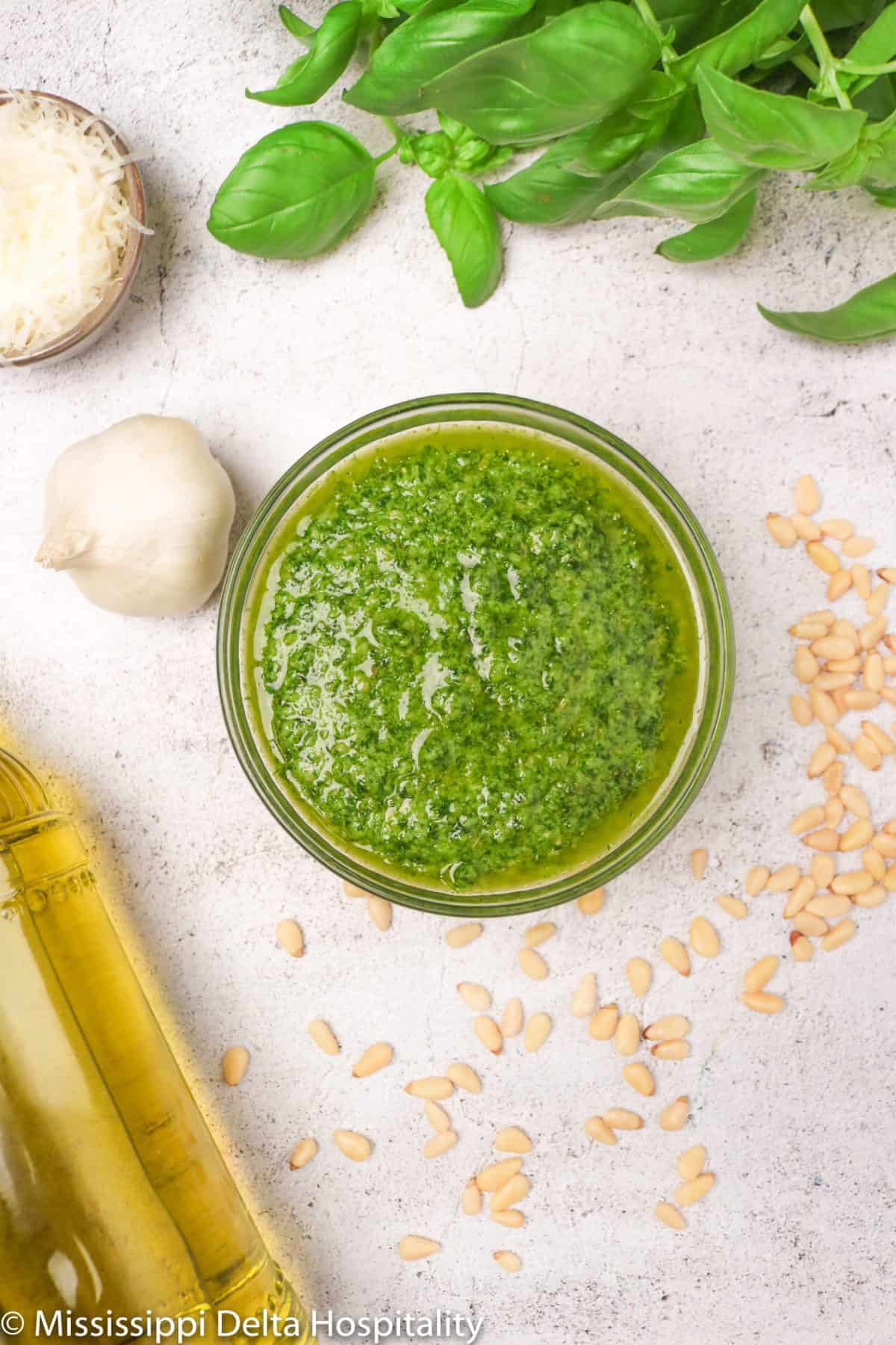 a small glass bowl full of basil pesto with a bottle of olive oil, a pod of garlic, a small bowl of grated parmesan cheese, a stack of basil leaves, and pine nuts on a concrete board.