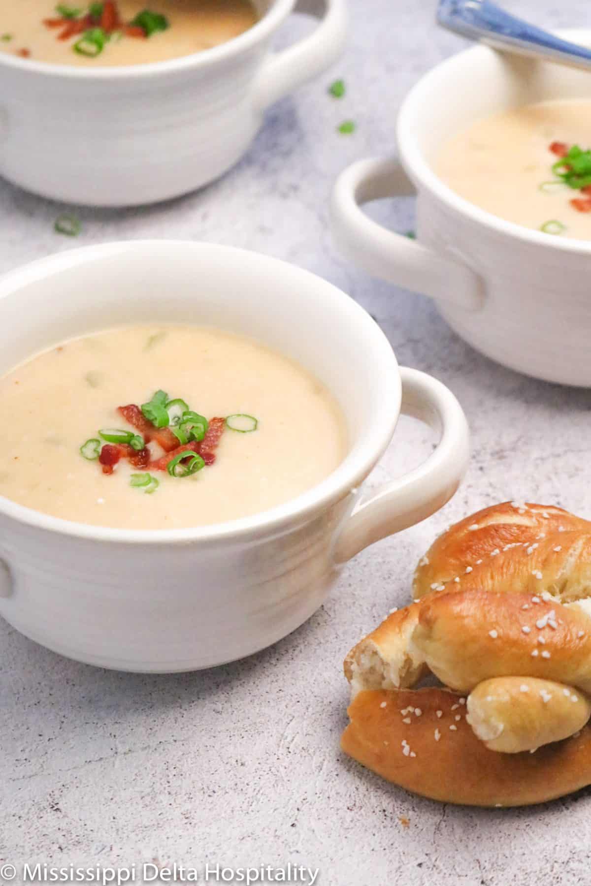 Three white farmhouse bowls of cheddar cheese soup with a pretzel on a concrete board
