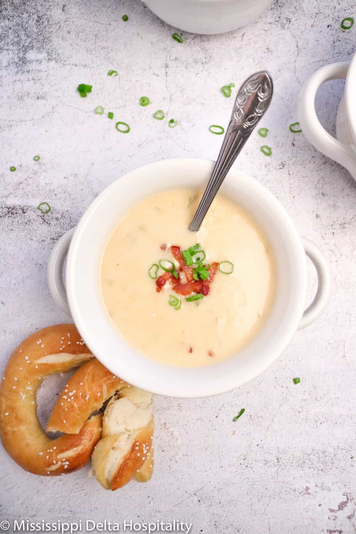a white farmhouse bowl full of cheddar cheese soup with a silver spoon in it and a pretzel.
