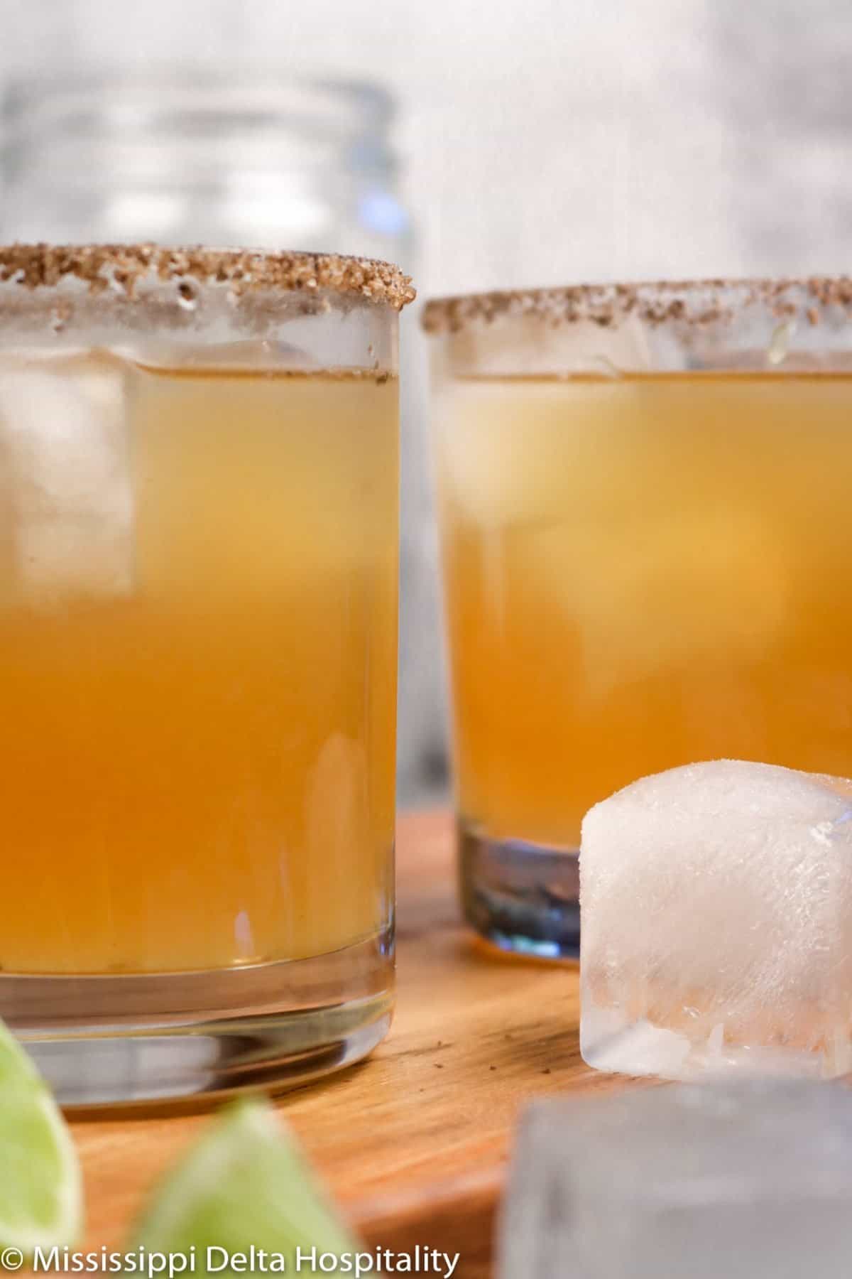 two glasses of margaritas on a wood board with two pieces of ice and two slices of lime.