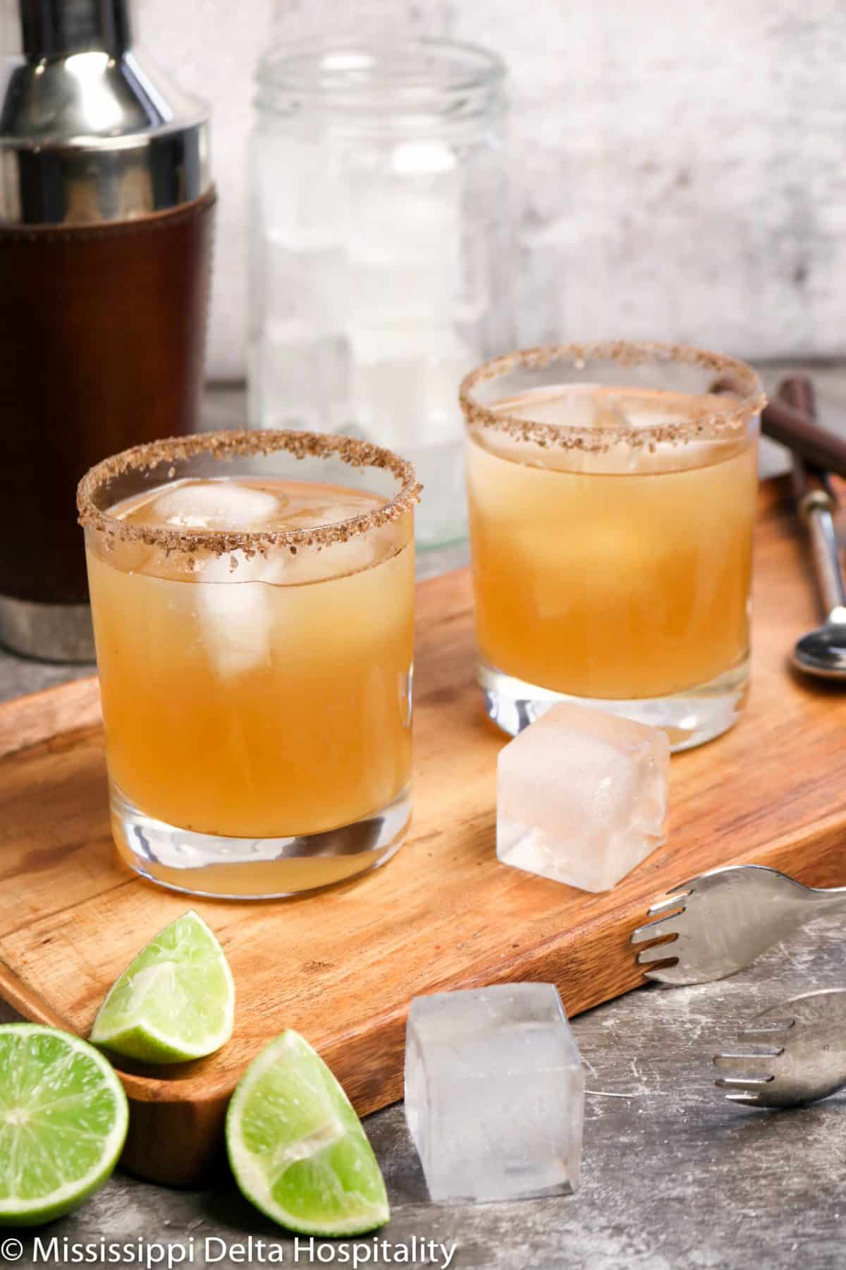 two glasses of margaritas sitting on a wood board with ice and slices of lime along with a cocktail shaker, a jar of ice, tongs, and a stirrer on a concrete board.