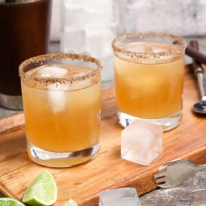 two glasses of margaritas sitting on a wood board with ice and slices of lime along with a cocktail shaker, a jar of ice, tongs, and a stirrer on a concrete board.