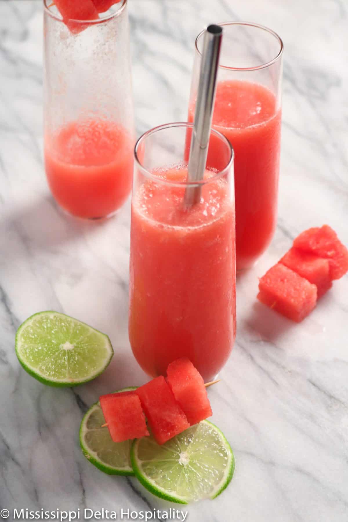 three glasses of watermelon slushie on a marble board with chunks of watermelon and slices of lime