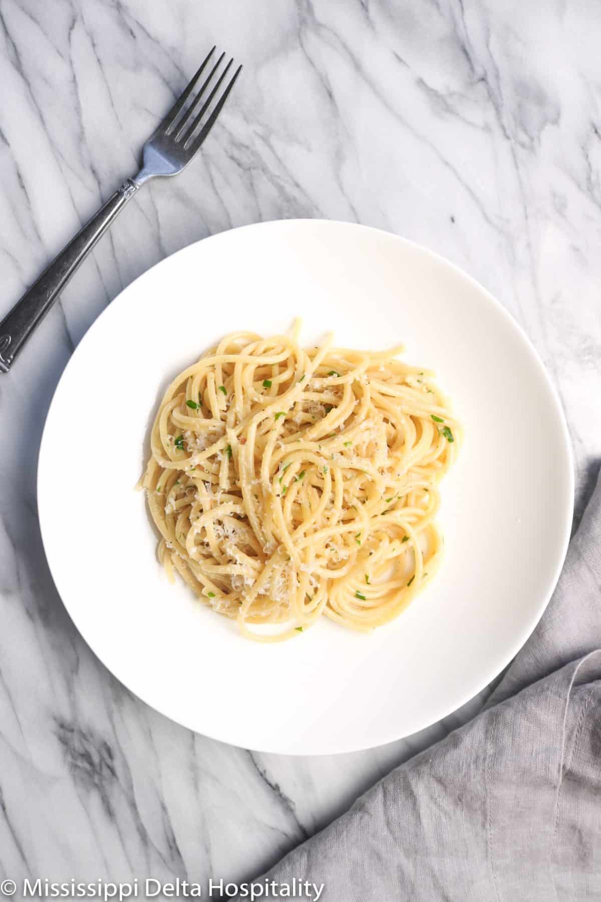 a white bowl with spaghetti aglio e olio on a marble board with a fork and a grey napkin