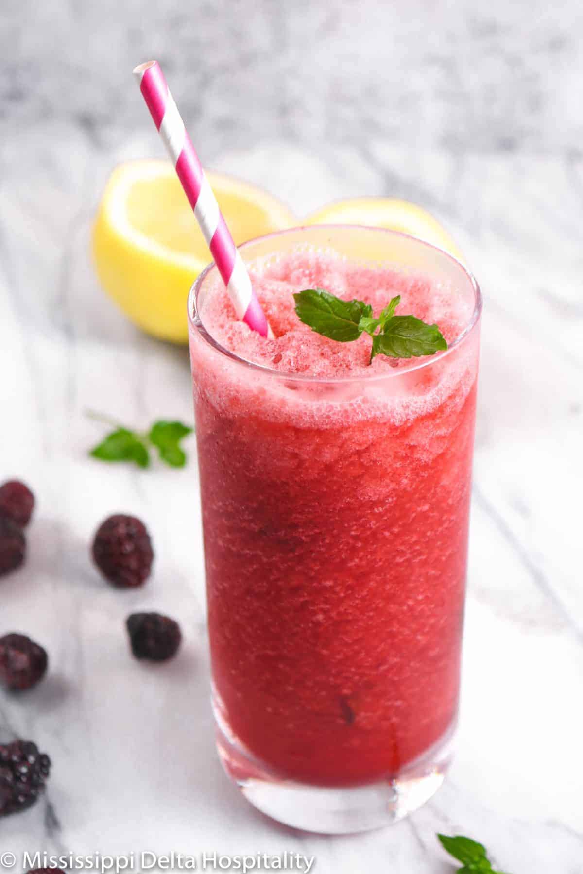 blackberry lemonade in a highball glass with mint, blackberries, and a lemon cut in half and a pink striped straw