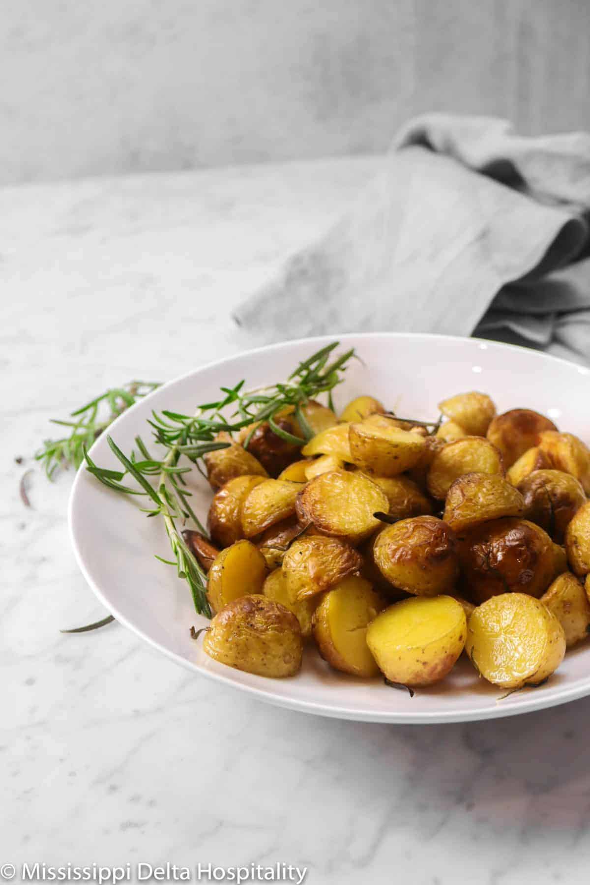 roasted potatoes in a white serving bowl with a sprig of rosemary on a marble board with a grey napkin