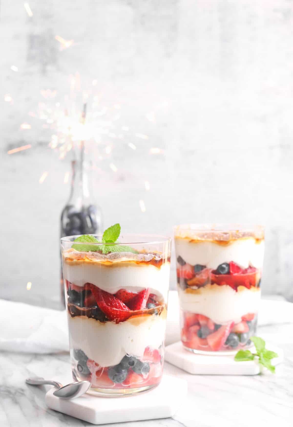 two glasses of berries and cream parfait with a jar of blueberries and a sparkler