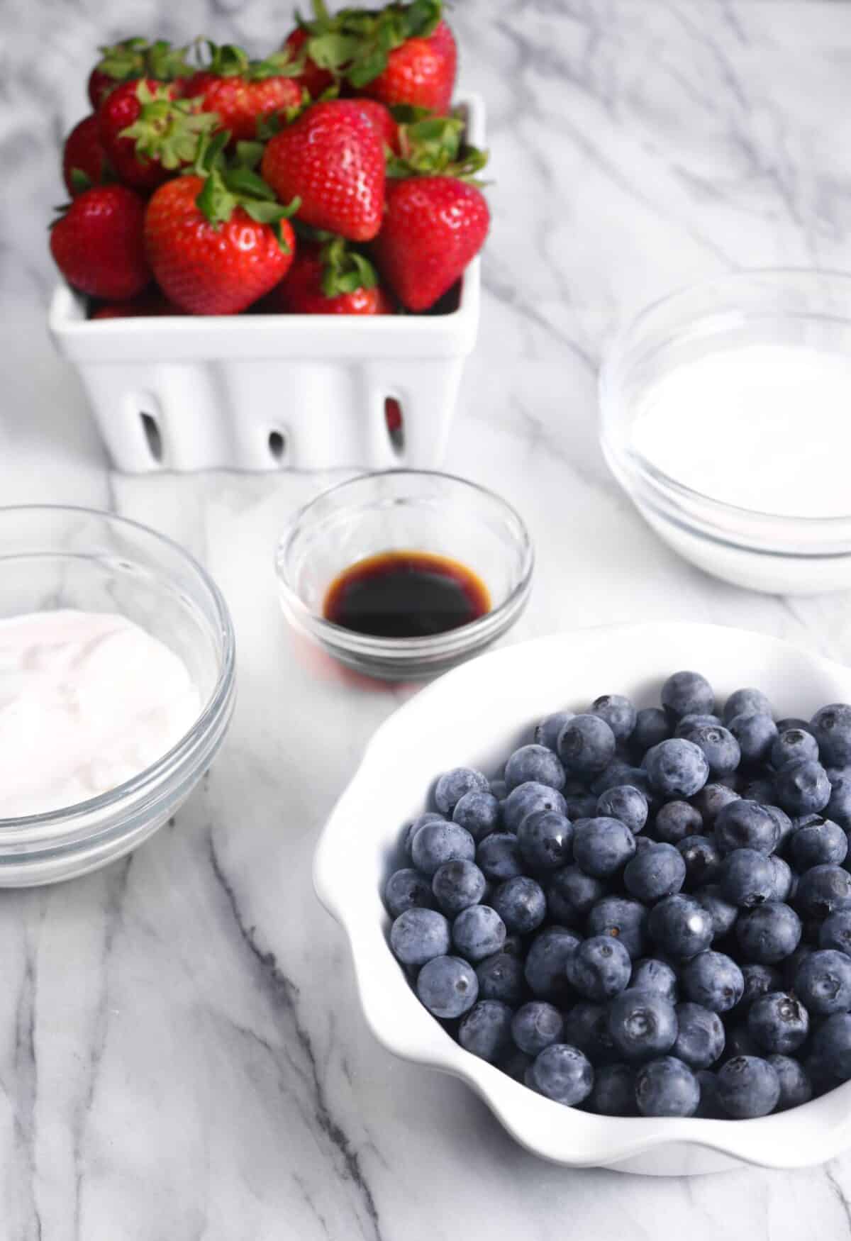 a bowl of blueberries, a basket of strawberries, and three bowls with heavy cream, vanilla, and sour cream on a marble board