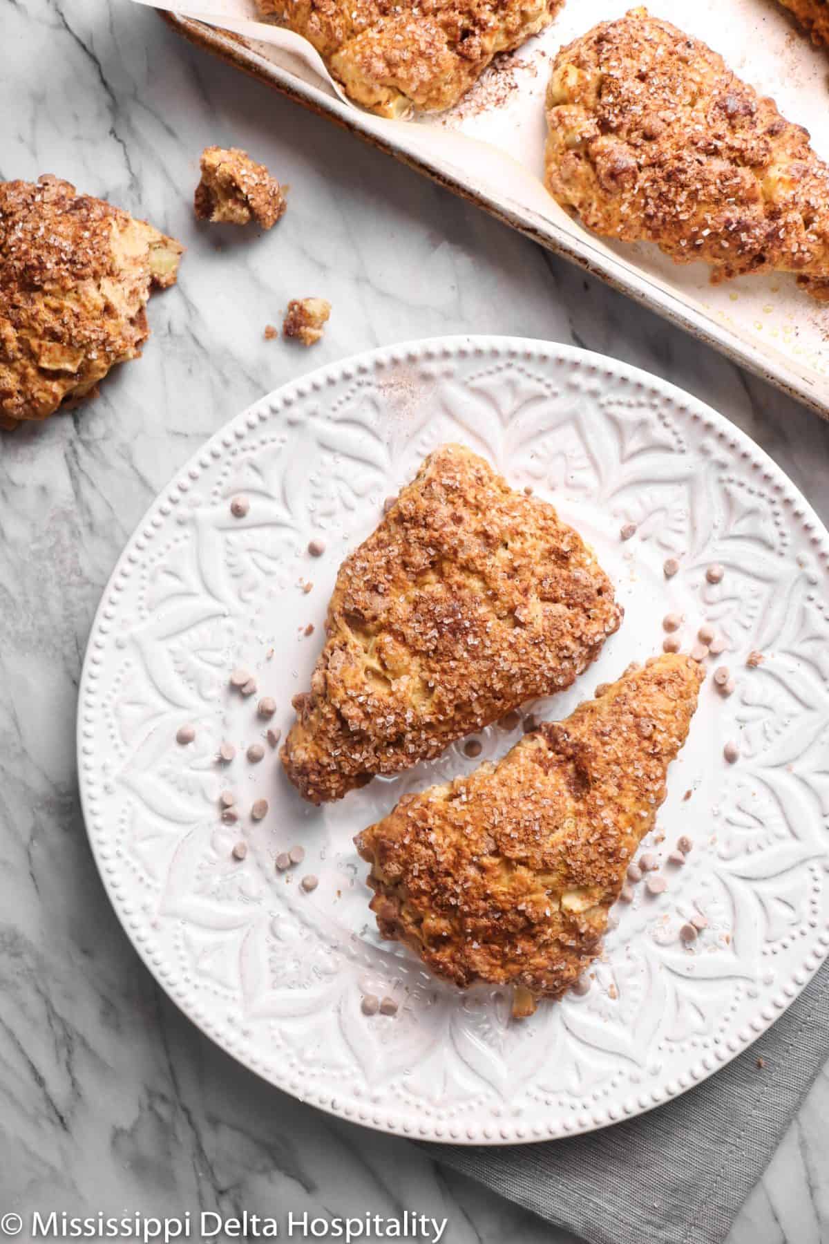a white plate with two apple cinnamon scones and a sheet pan with scones on a marble board