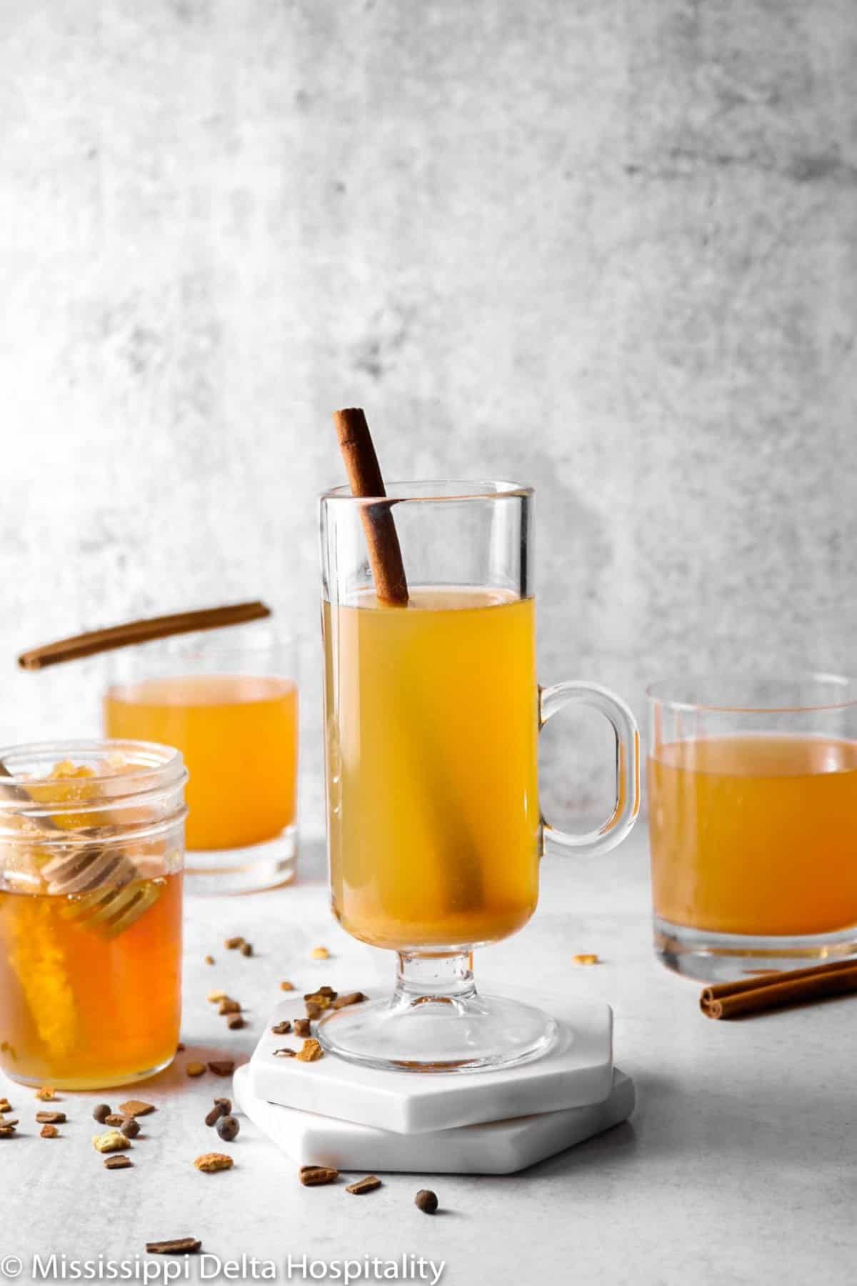 three glasses of spple cider on a concrete board with cinnamon sticks and a jar of honey