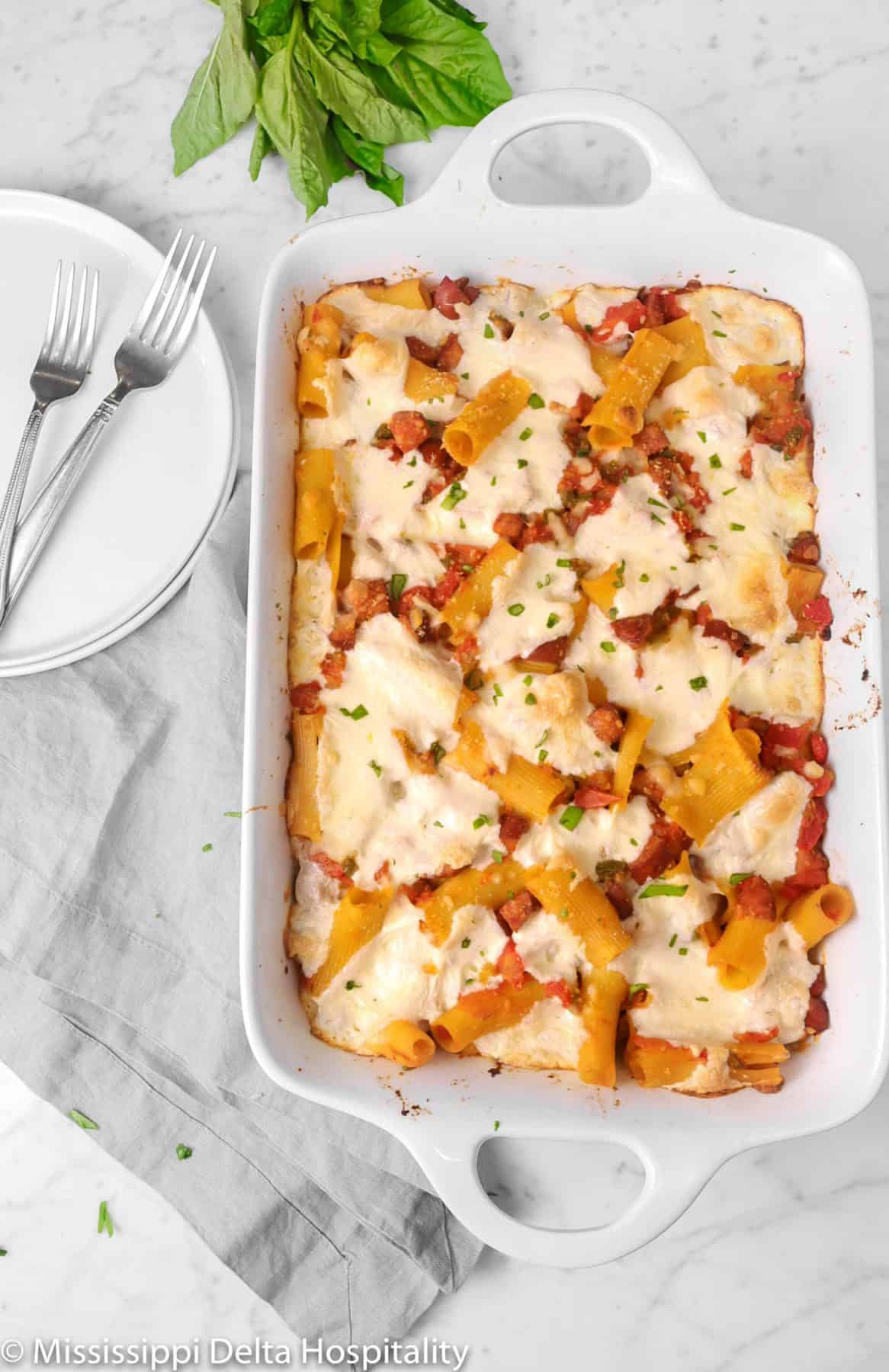 baked rigatoni on a marble board with two plates, two forks, a grey napkin, and basil leaves