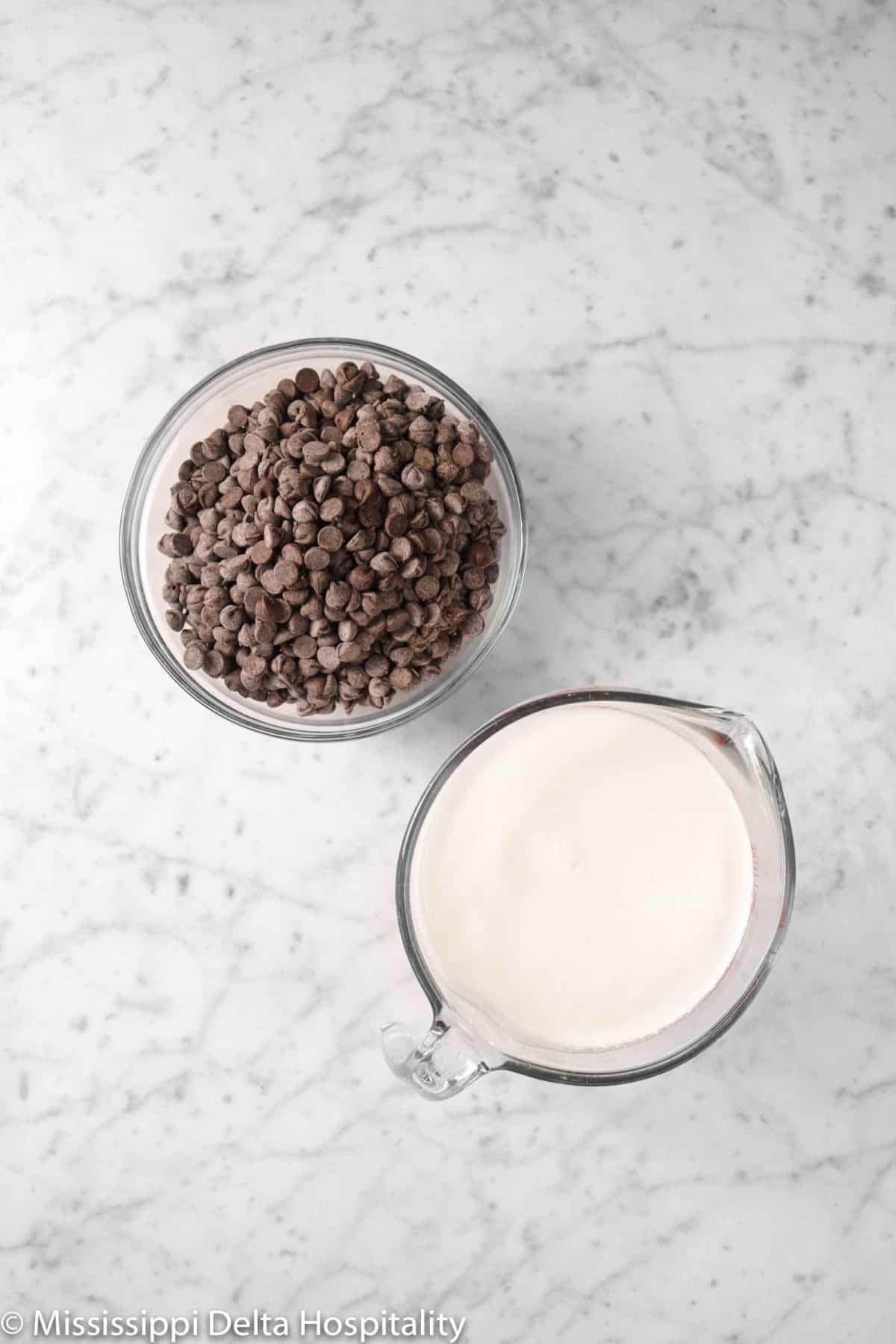 a measuring cup of heavy cream and a bowl of chocolate chips on a marble board