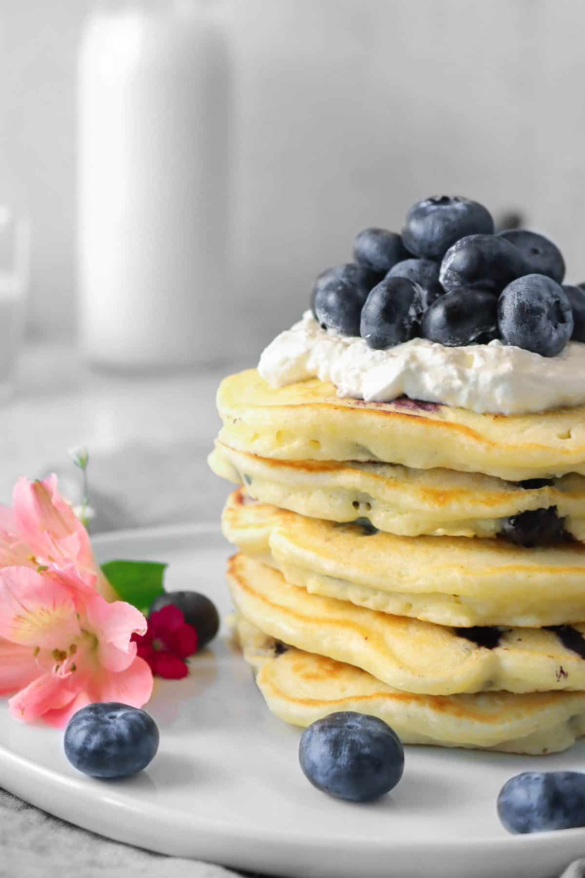 a stack of blueberry pancakes with whipped cream, fresh blueberries, and flowers on a white plate with a jar of milk in the background