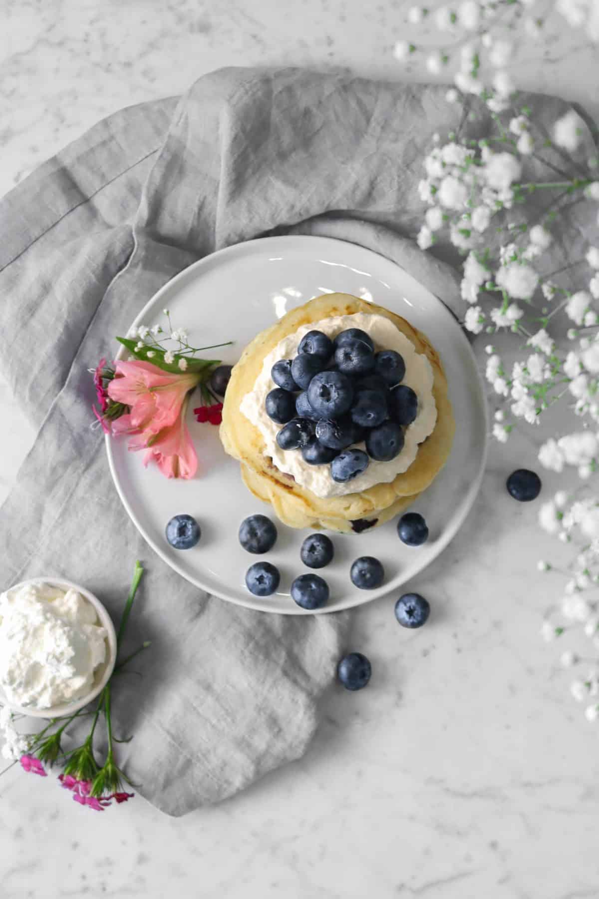 blueberry pancakes on a white plate with whipped cream, a grey napkin, and flowers