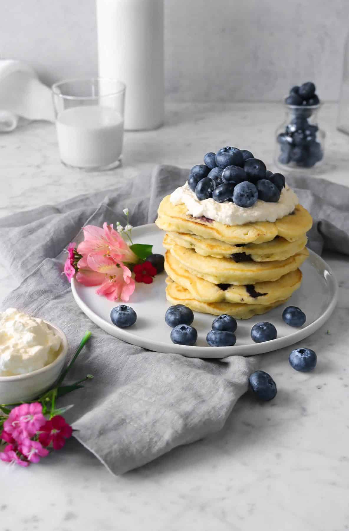 a stack of blueberry pancakes on a white plate with blueberries, a grey napkin and a glass of milk and flowers