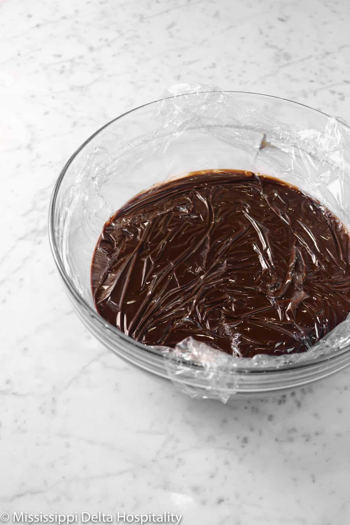 melted chocolate in a glass bowl with plastic wrap over it on a marble table