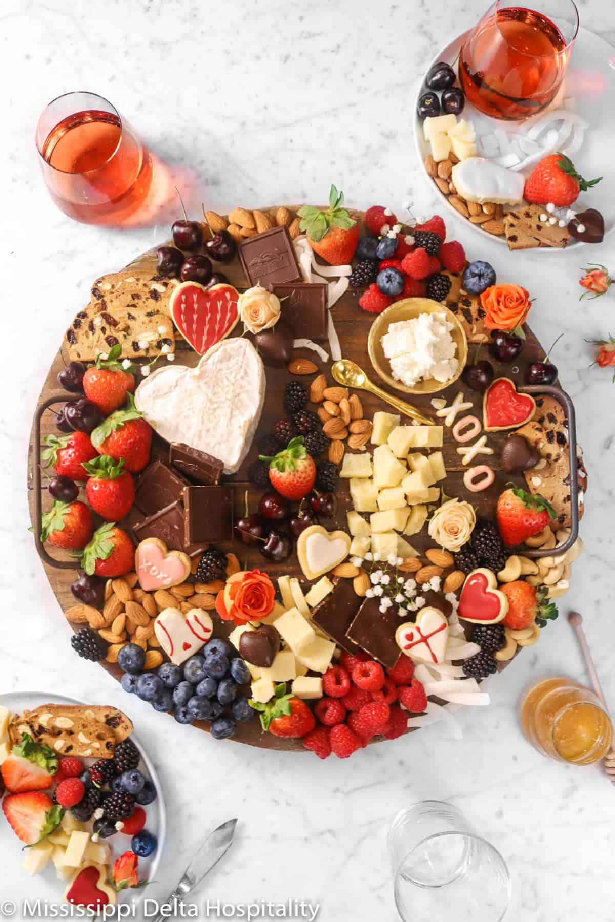 wood serving board with cheese, chocolates, heart shaped cookies, berries, and fresh flowers on a marble board with two plates and glasses of rosé and a jar of honey