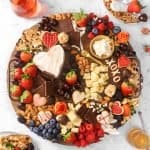 wood serving board with cheese, chocolates, heart shaped cookies, berries, and fresh flowers on a marble board with two plates and glasses of rosé and a jar of honey