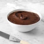 a bowl of chocolate frosting on a marble counter with a white napkin and a frosting spreader