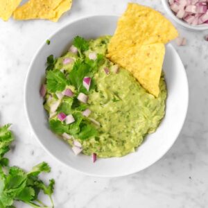 A bowl of guacamole on a marble table with cilantro, red onions, and corn chips