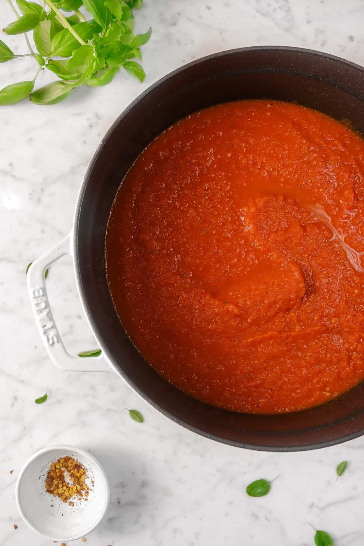 a pot of tomato sauce on a marble counter with basil and red pepper