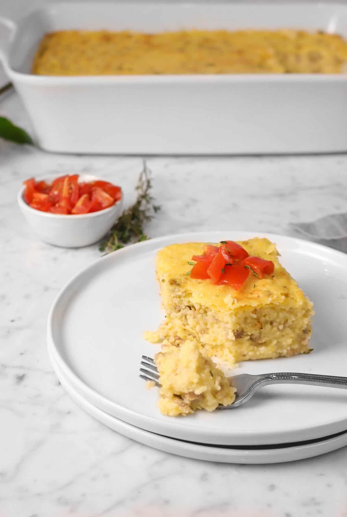 a slice of of grits casserole on a white plate with tomatoes and a bite taken out of it