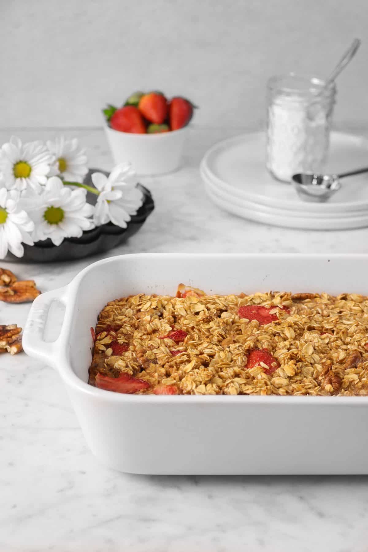 strawberry oatmeal bake in a casserole dish with flowers, strawberries and plates in the background