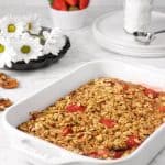 strawberry oatmeal bake in a white casserole dish on a marble counter with flowers, strawberries, white plates, and powdered sugar in the background