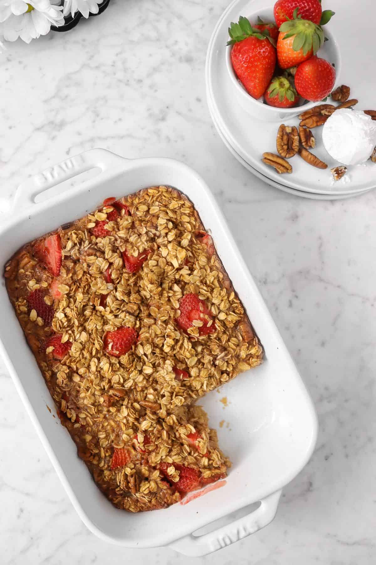 strawberry oatmeal bake with a piece cut out of it on a marble counter with white plates, strawberries, powdered sugar, and pecans
