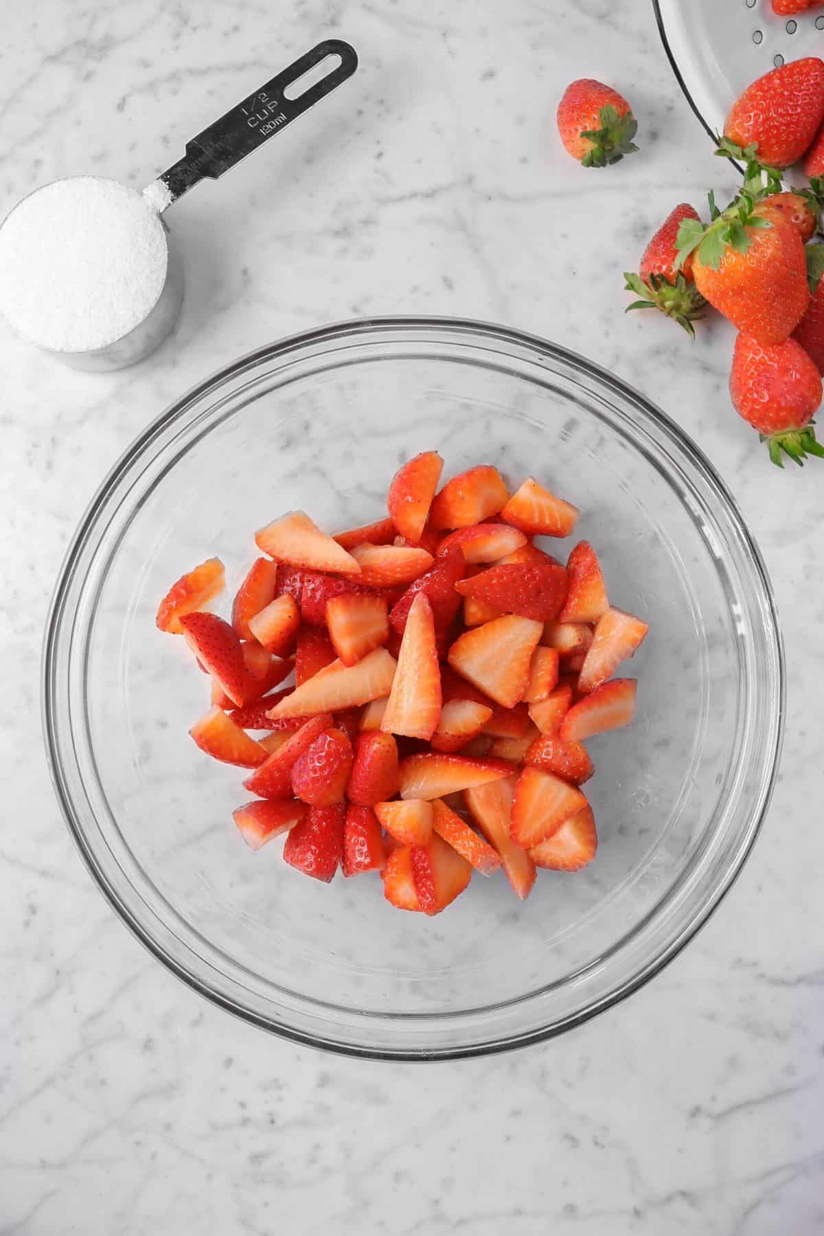 strawberries chopped in a glass bowl with a cup of sugar and extra strawberries