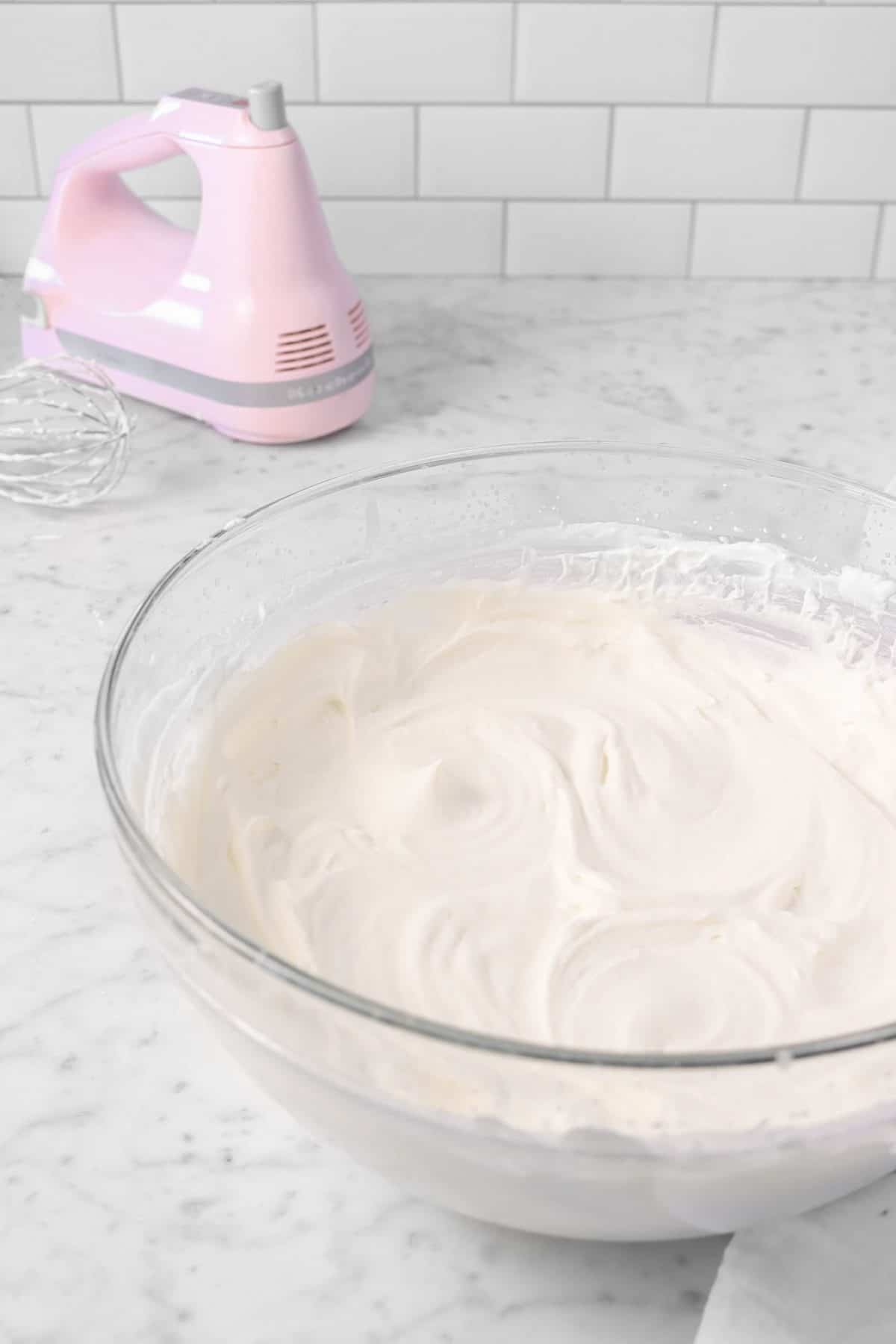 chantilly cream in a glass bowl with a pink hand mixer on a marble counter