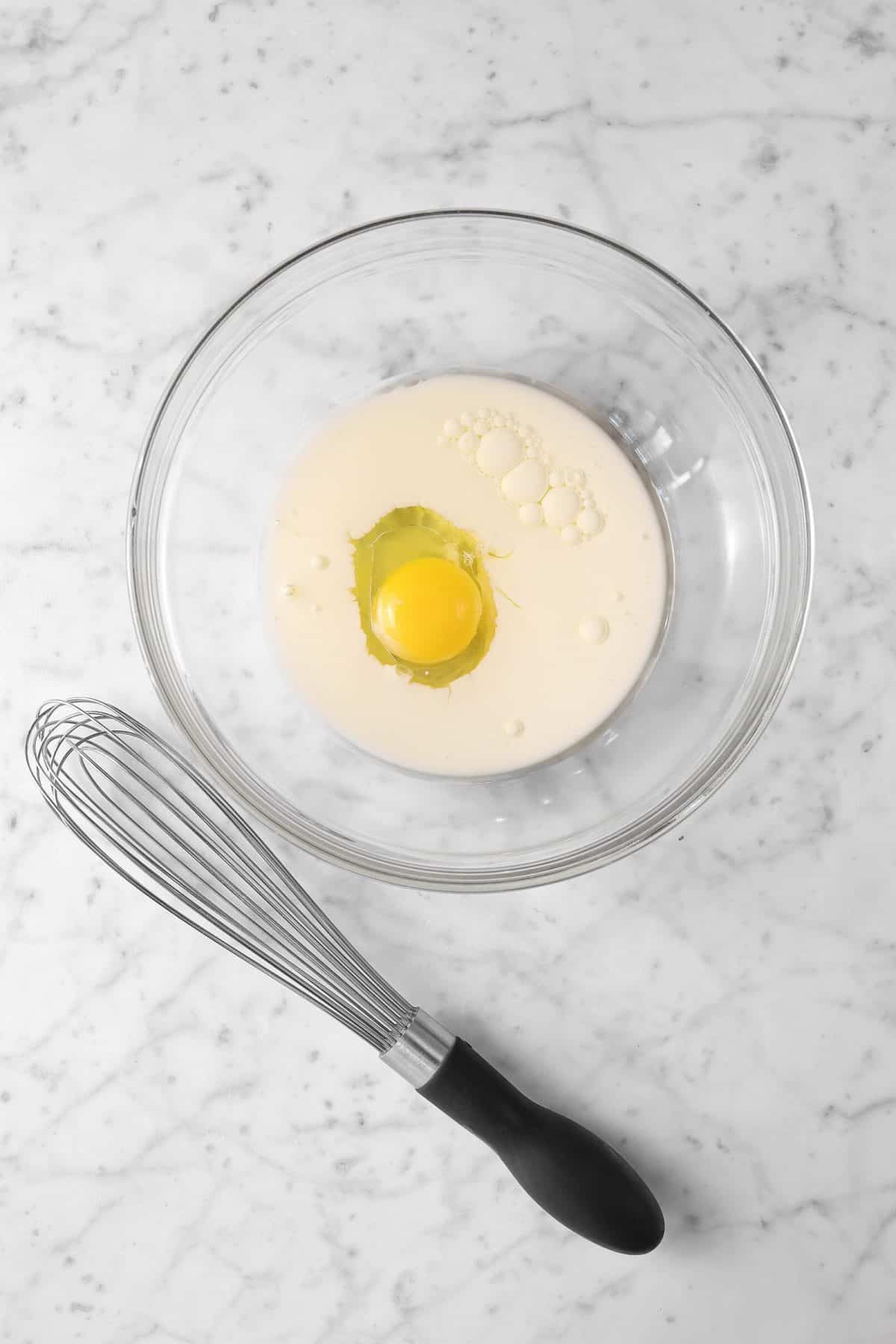 egg, milk, and oil in a glass bowl with a whisk on a marble counter
