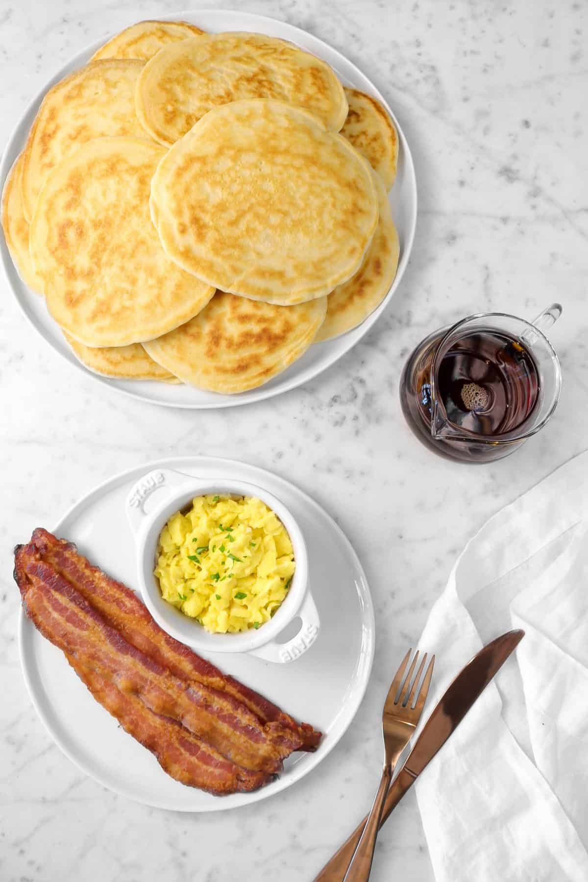 sourdough pancakes on a white plate with a plate of bacon and eggs and small glass of maple syrup