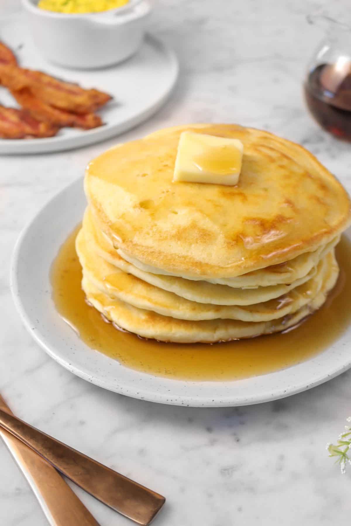 a stack of sourdough pancakes with maple syrup on a white plate with a plate of bacon and eggs in the background