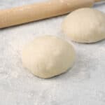 two boules of pizza dough on a floured marble surface with a wood rolling pin