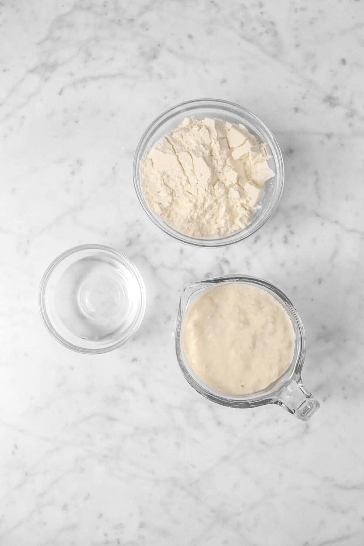 sourdough starter, flour, and water in glass bowls on a marble counter