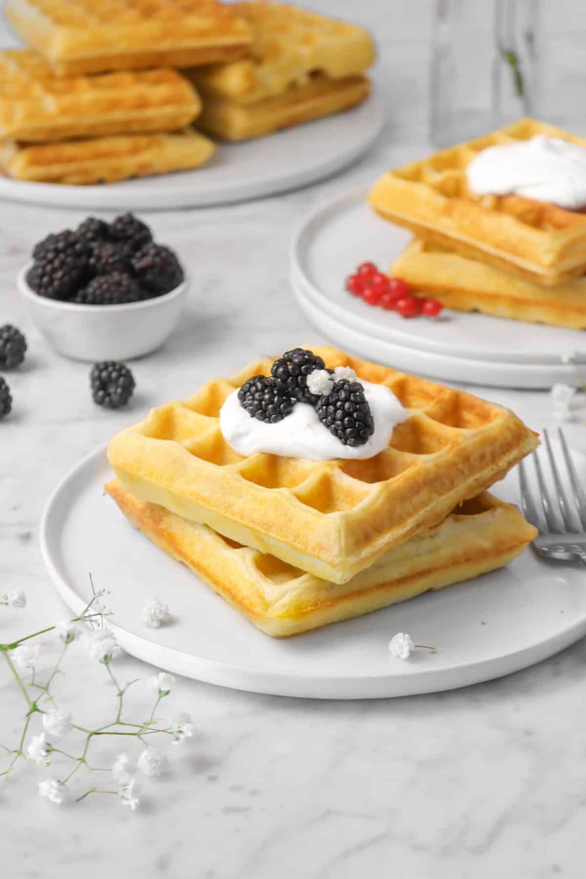 two waffles on a white plate with whipped cream and blackberries and white flowers with two other plates of waffles behind it