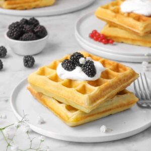 two waffles on a white plate with whipped cream and blackberries and white flowers with two other plates of waffles behind it