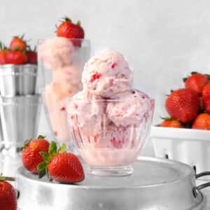 four scoops of strawberry ice cream in a glass bowl on a stainless steel baking pan with fresh strawberries and a milk shake glass with four scoops of strawberry ice cream in the background