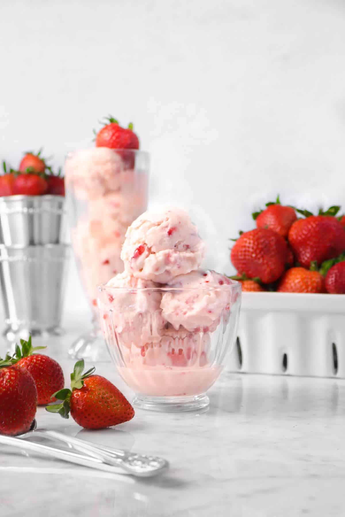 four scoops of strawberry ice cream in a glass bowl with fresh strawberries, two mint julep cups, and a milkshake glass