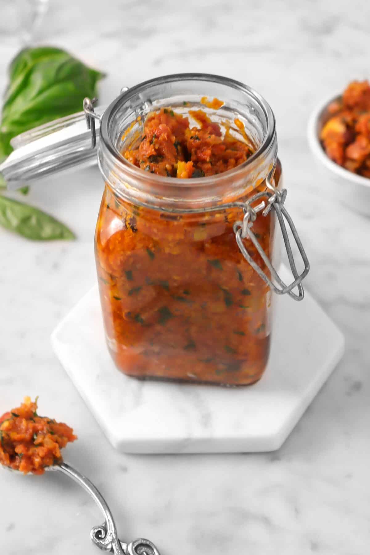 tomato pesto in a glass jar with basil leaves and a spoonful of pesto on a marble counter