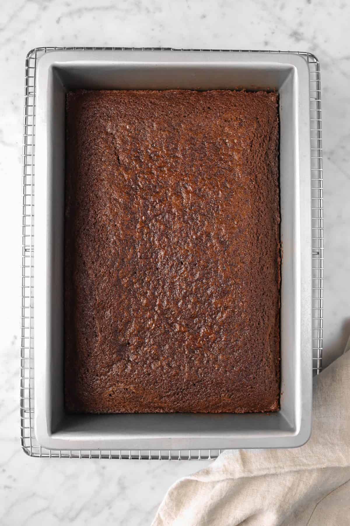chocolate cake in a large rectangular cake pan on a wire cooling rack with a brown napkin