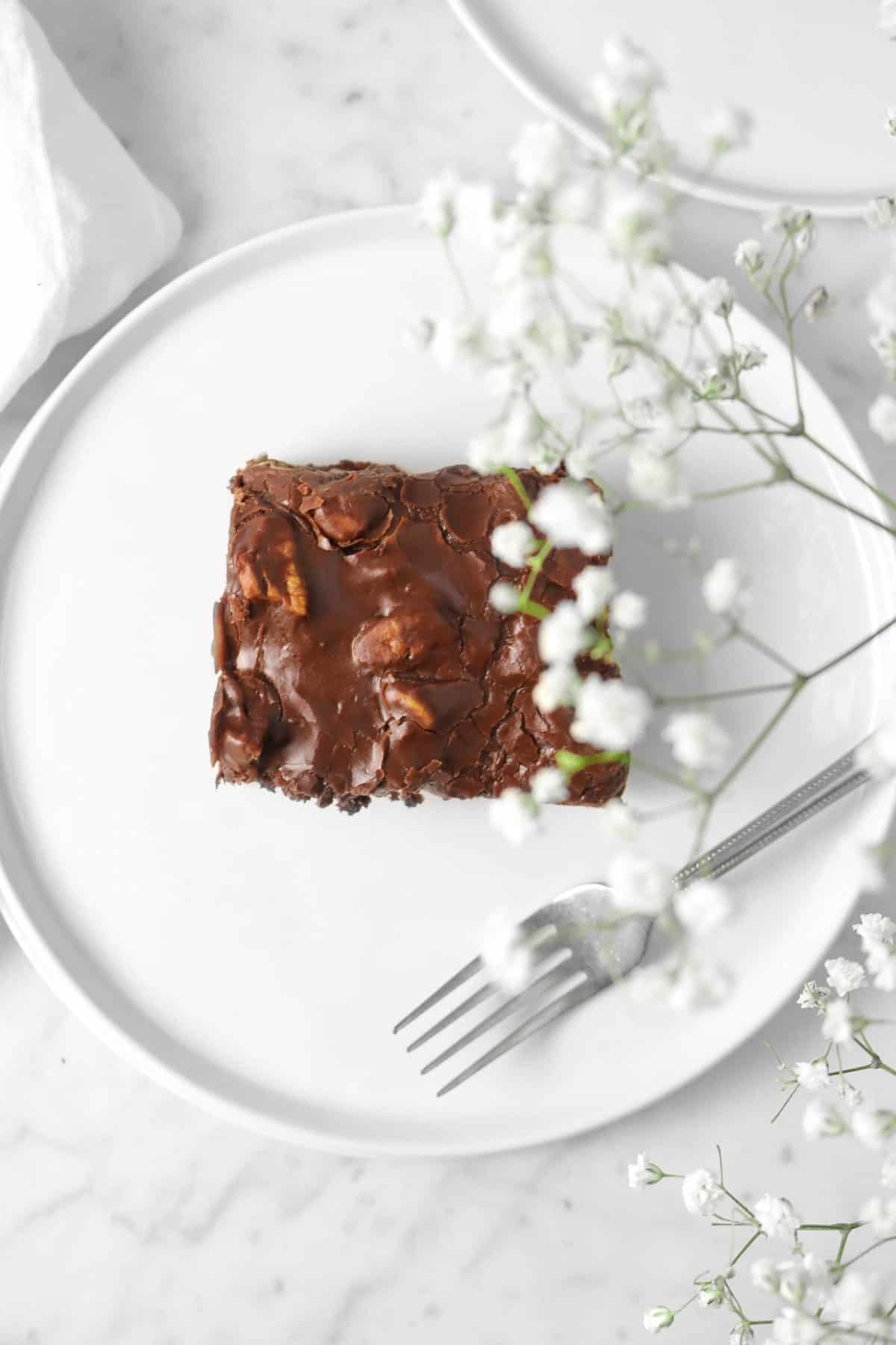 an overhead shot of a square slice of texas chocolate sheet on a white plate with a fork and white flowers