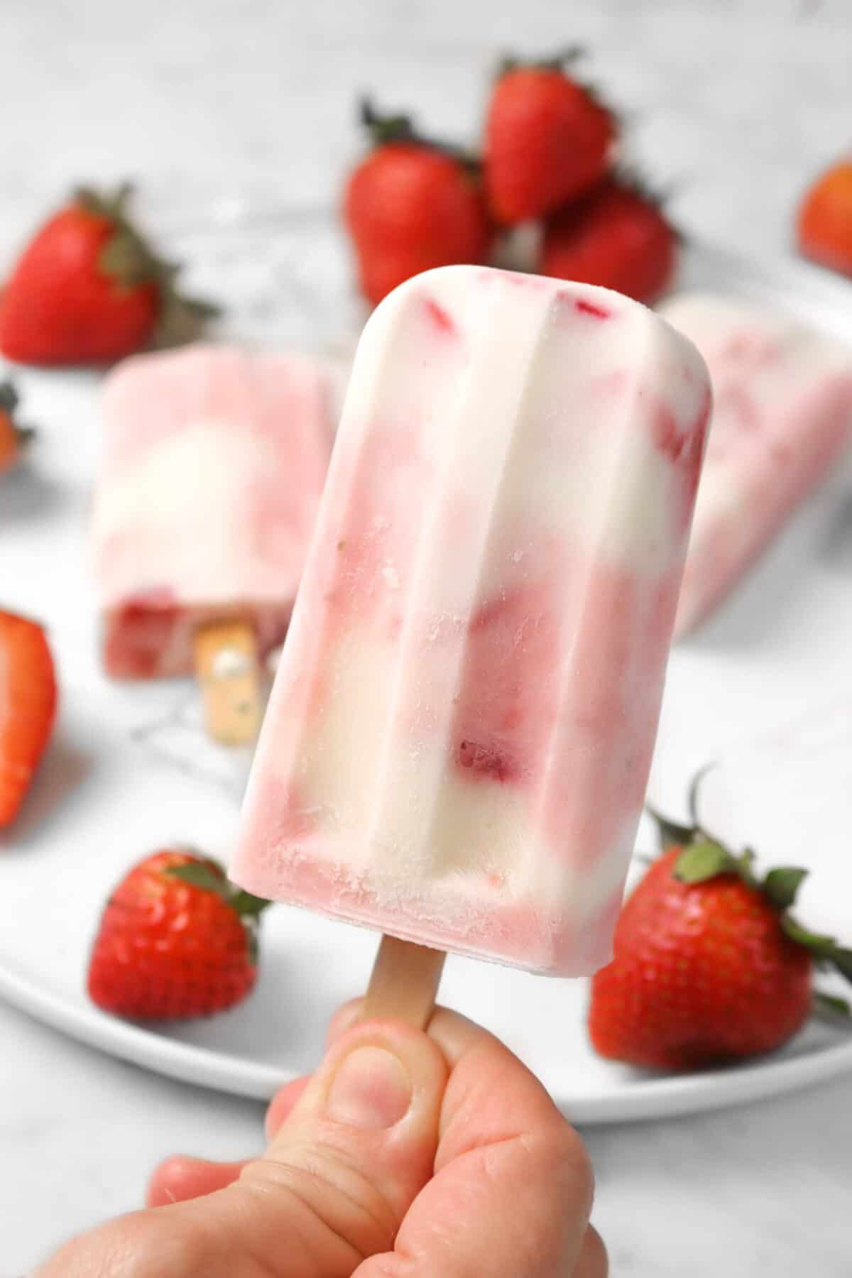 hand holding strawberry yogurt popsicle with two popsicles in the background with strawberries
