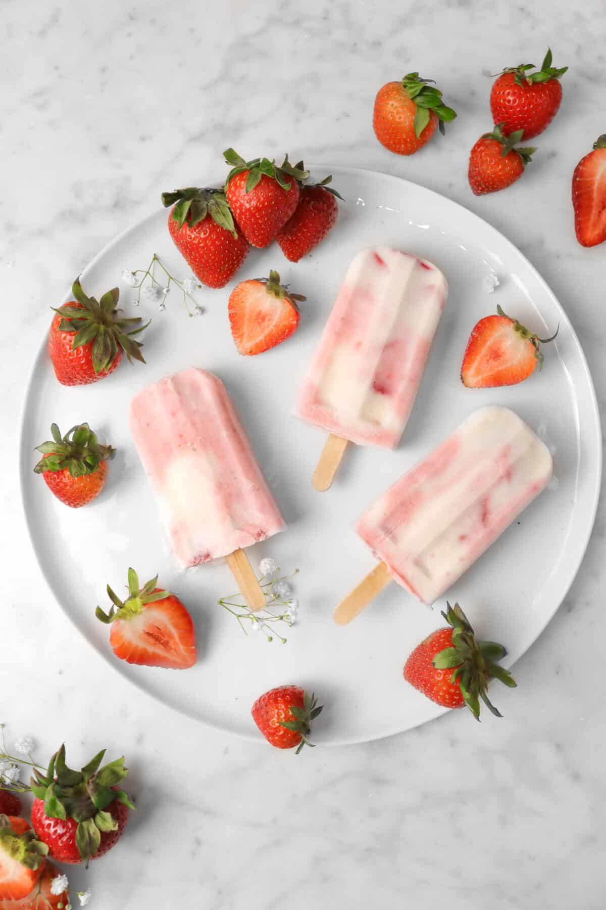 overhead shot of three strawberry yogurt popsicles on a white plate with strawberries and fresh flowers