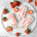 overhead shot of three strawberry yogurt popsicles on a white plate with strawberries and fresh flowers