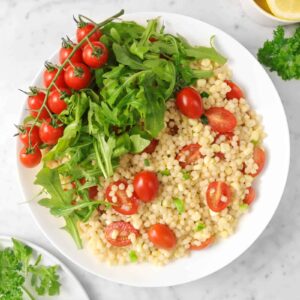 couscous salad in a white bowl with arugula, tomato on the vine, herbs, lemon slices, and oil in a bowl